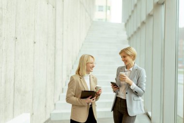 Ofis koridorunda dijital tablet ve cep telefonuyla yürüyen iki iş kadını.