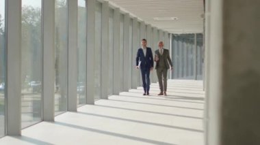 Yakışıklı iş adamı ve genç meslektaşı ofis koridorunda mali raporları tartışıyorlar.