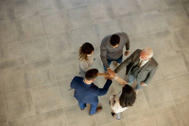 Group of young and senior business people come together in an office hallway, standing in a circle with their hands joined. They are dressed professionally, reflecting their business acumen and status. Their teamwork and collaboration symbolize the u