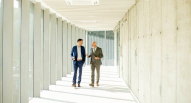 Young and a senior businessman walk down an office hallway, deep in conversation. They are both dressed professionally, reflecting their business acumen and status. They are navigating the complexities of the corporate world, finding solutions and ma