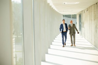 Young and a senior businessman walk down an office hallway, deep in conversation. They are both dressed professionally, reflecting their business acumen and status. They are navigating the complexities of the corporate world, finding solutions and ma