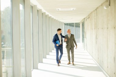 Young and a senior businessman walk down an office hallway, deep in conversation. They are both dressed professionally, reflecting their business acumen and status. They are navigating the complexities of the corporate world, finding solutions and ma