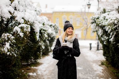 Güzel, genç bir bayan, sıcak kıyafetler içinde, karın içinde kahve fincanıyla eğleniyor.