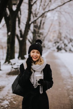 A young woman wearing warm winter clothes and a knit hat smiles happily as she stands in the snow and using mobile phone whil holding cofee cup