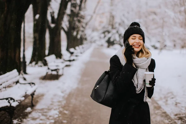 一位身穿温暖冬衣 头戴针织帽子的年轻女子 站在雪地里 拿着咖啡杯 用手机欢快地微笑着 — 图库照片