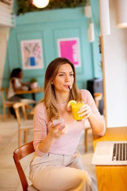 Laptop üzerinde çalışırken cep telefonu kullanan ve kafede taze portakal suyu içen güzel bir kadın.