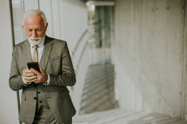 A senior business man stands in an office hallway, focused on his mobile phone. He is dressed in formal attire, exuding confidence and professionalism