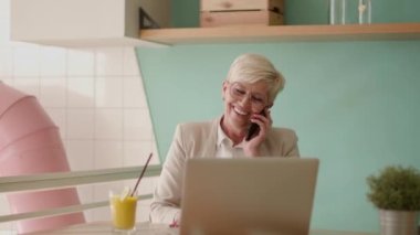 Üst düzey kadın laptopta çalışırken cep telefonu kullanıyor ve kafede taze portakal suyu içiyor.