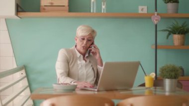 Üst düzey kadın laptopta çalışırken cep telefonu kullanıyor ve kafede sağlıklı bir kahvaltı yapıyor.