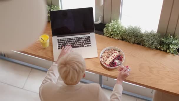 老年妇女一边在笔记本电脑上工作一边用手机 在咖啡店里吃着健康的早餐 — 图库视频影像