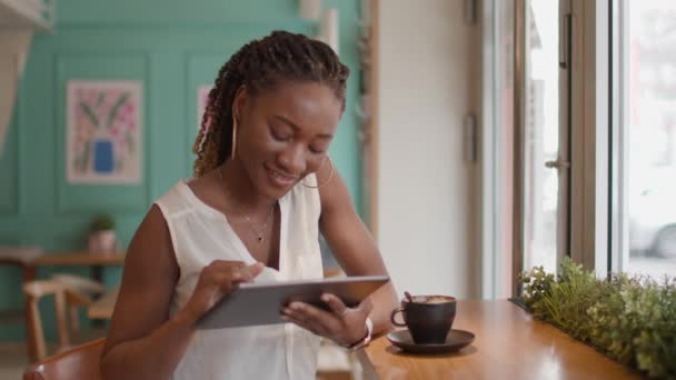 漂亮的黑人女人一边喝咖啡 一边看着咖啡店里的数码平板电脑 — 图库视频影像