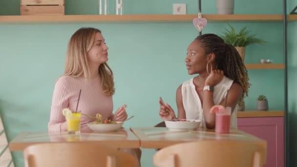 两个年轻貌美的黑人和高加索女人在咖啡馆里玩得很开心 喝着新鲜的果汁 吃着健康的早餐 — 图库视频影像
