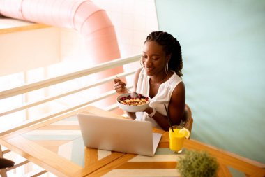 Güzel siyah kadın kafede dizüstü bilgisayarla çalışırken sağlıklı bir kahvaltı yapıyor.