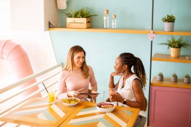İki güzel siyah ve beyaz kadın iyi vakit geçiriyor, taze meyve suyu içiyor ve kafede sağlıklı bir kahvaltı yapıyor.
