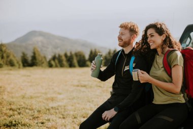 Smiling young couple preparing hiking adventure with backpacks by terrain vehicle