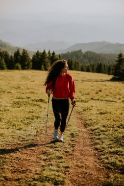 Sırt çantasıyla yeşil tepelerde yürüyen gülümseyen genç kadın.