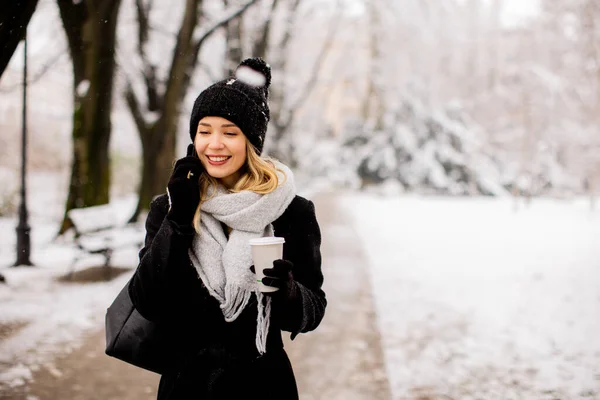 一位身穿温暖冬衣 头戴针织帽子的年轻女子 站在雪地里 拿着咖啡杯 用手机欢快地微笑着 — 图库照片