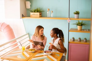 İki güzel siyah ve beyaz kadın iyi vakit geçiriyor, taze meyve suyu içiyor ve kafede sağlıklı bir kahvaltı yapıyor.