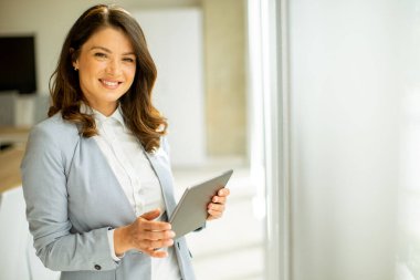Ofis penceresinin yanında dijital tablet üzerinde çalışan güzel genç bir kadın.