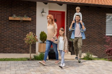 Family with a mother, father, son and daughter walking with  abaggage outside on the front porch of a brick house clipart