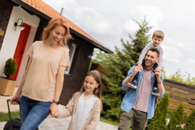 Bir anne, baba, oğul ve kızı olan aile tuğladan bir evin verandasında abagage ile yürüyor.
