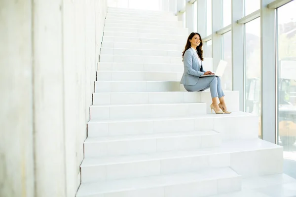 若いビジネスマンの女性がオフィスの廊下の階段に座ってノートパソコンで働いています — ストック写真