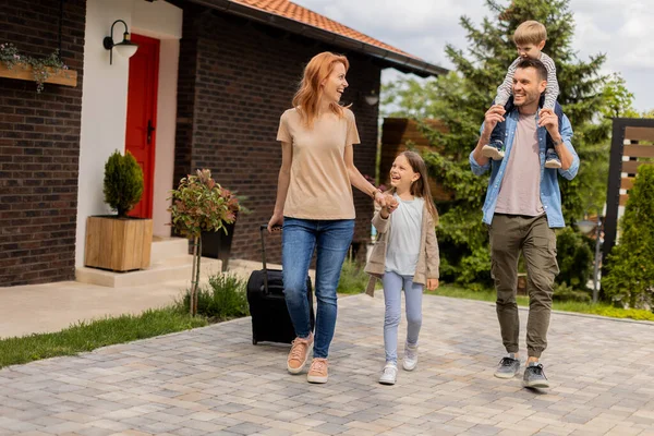 Bir anne, baba, oğul ve kızı olan aile tuğladan bir evin verandasında abagage ile yürüyor.