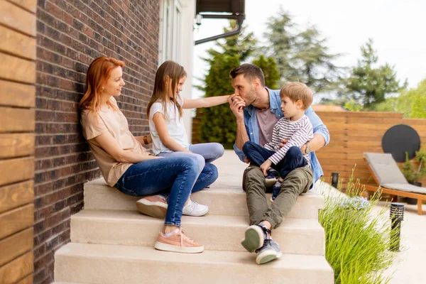 Bir anne, baba, oğul ve kızı olan aile dışarıda tuğladan bir evin verandasında oturuyor.