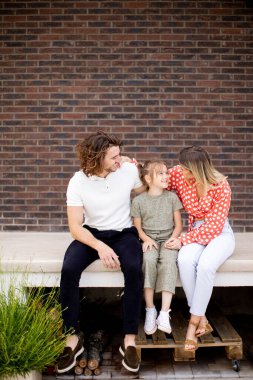 Bir anne, baba ve kızı olan aile tuğladan bir evin verandasının önünde oturuyor.
