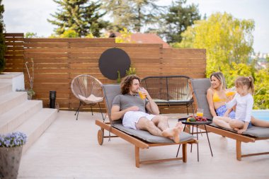 Happy family with a mother, father and daughter sitting on the deck chairs by the swimming pool