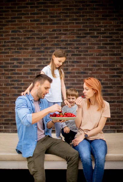 Family with a mother, father, son and daughter sitting outside on a steps of a front porch of a brick house and eating strawberries