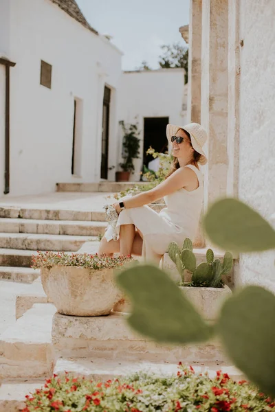 A young woman in a white dress and  hat relaxing on stairs on a sunny day during tourist visit in Alberobello, Italy