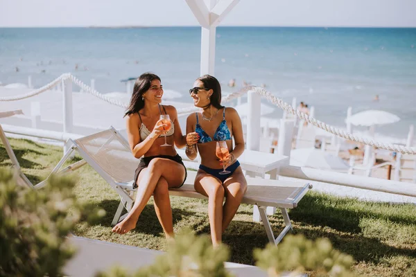 カクテルを飲んでビーチで休暇を楽しんでいるビキニの2人の笑顔の若い女性 — ストック写真