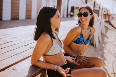 Bikinili iki gülümseyen genç kadın sahilde oturup tatilin tadını çıkarıyor.