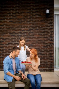 Family with a mother, father and daughter sitting outside on a steps of a front porch of a brick house and eating strawberries