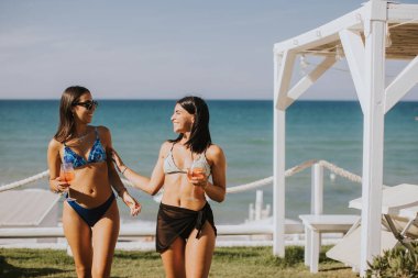 İki gülümseyen bikinili genç kadın kokteyl içerken sahilde tatilin tadını çıkarıyor.