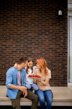 Family with a mother, father and daughter sitting outside on a steps of a front porch of a brick house and eating strawberries