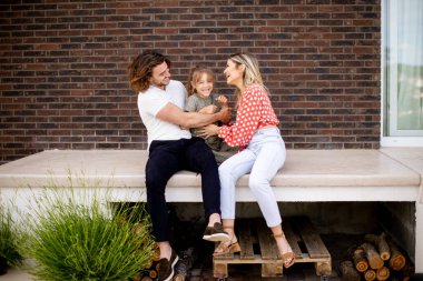 Bir anne, baba ve kızı olan aile tuğladan bir evin verandasının önünde oturuyor.