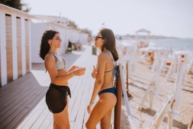 Bikinili iki gülümseyen genç kadın kulübelerin yanında duruyor ve sahilde tatilin tadını çıkarıyorlar.