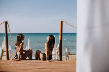 Bikinili iki gülümseyen genç kadın sahilde oturup tatilin tadını çıkarıyor.