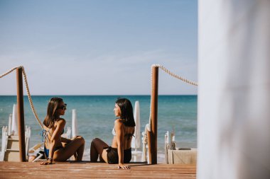 Bikinili iki gülümseyen genç kadın sahilde oturup tatilin tadını çıkarıyor.