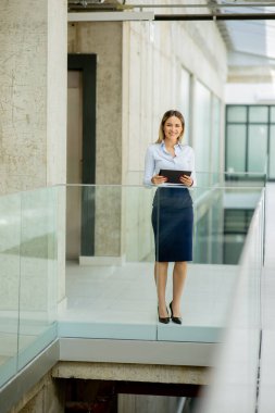 Modern ofis duvarında dijital tabletli genç iş kadını duruyor.