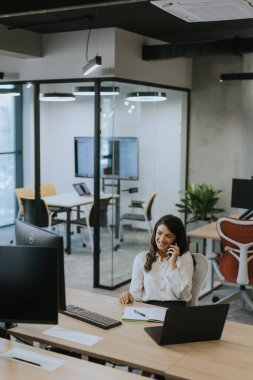 Modern ofiste dizüstü bilgisayarla çalışırken cep telefonu kullanan genç ve güzel bir iş kadını.