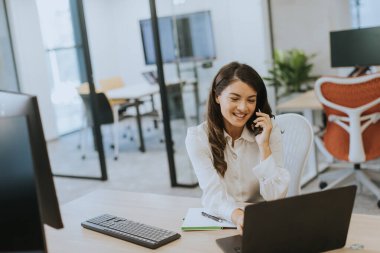 Modern ofiste dizüstü bilgisayarla çalışırken cep telefonu kullanan genç ve güzel bir iş kadını.