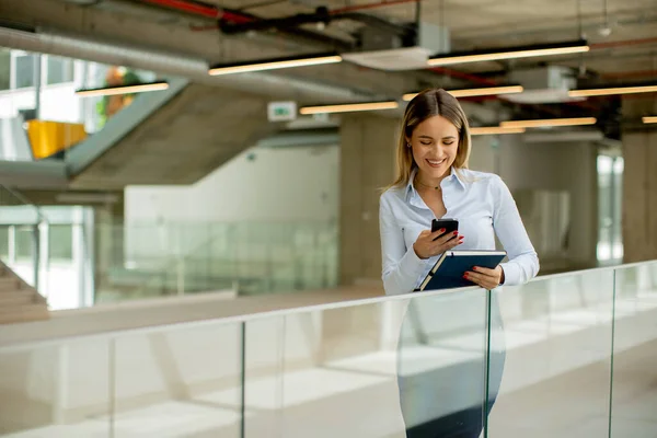 Young business woman with notebook using mobile phone and standing in the modern office wallway