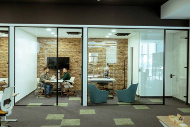 İki profesyonel, yeşil ve çağdaş tasarım unsurlarıyla çevrili bir ofis salonunda rahatça otururken sohbet ediyorlar.