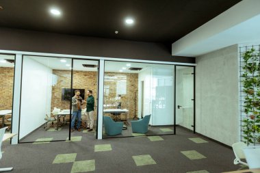 İki profesyonel, yeşil ve çağdaş tasarım unsurlarıyla çevrili bir ofis salonunda rahatça dururken sohbet ediyorlar.