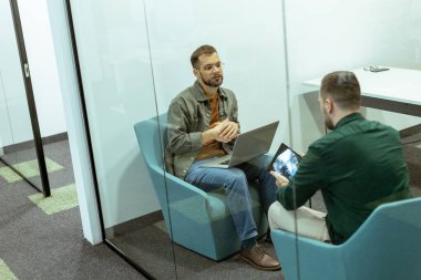 İki profesyonel, yeşil ve çağdaş tasarım unsurlarıyla çevrili bir ofis salonunda rahatça otururken sohbet ediyorlar.