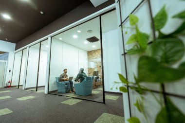 İki profesyonel, yeşil ve çağdaş tasarım unsurlarıyla çevrili bir ofis salonunda rahatça otururken sohbet ediyorlar.