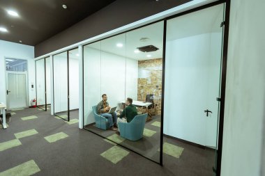 İki profesyonel, yeşil ve çağdaş tasarım unsurlarıyla çevrili bir ofis salonunda rahatça otururken sohbet ediyorlar.